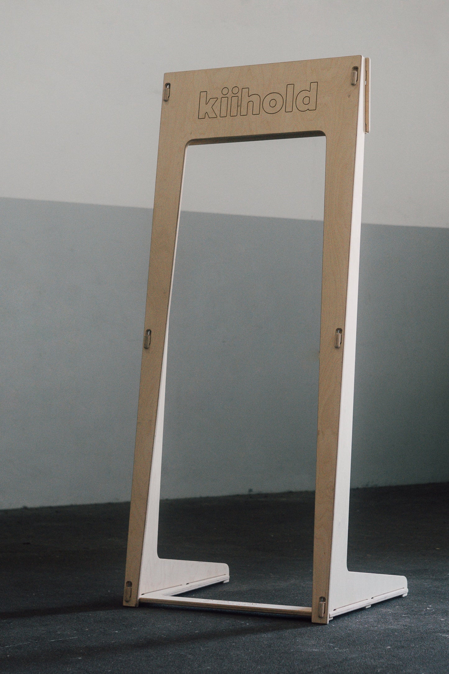 Freestanding wooden hanger with 'Kiihold' branding on a gray floor against a gray wall

