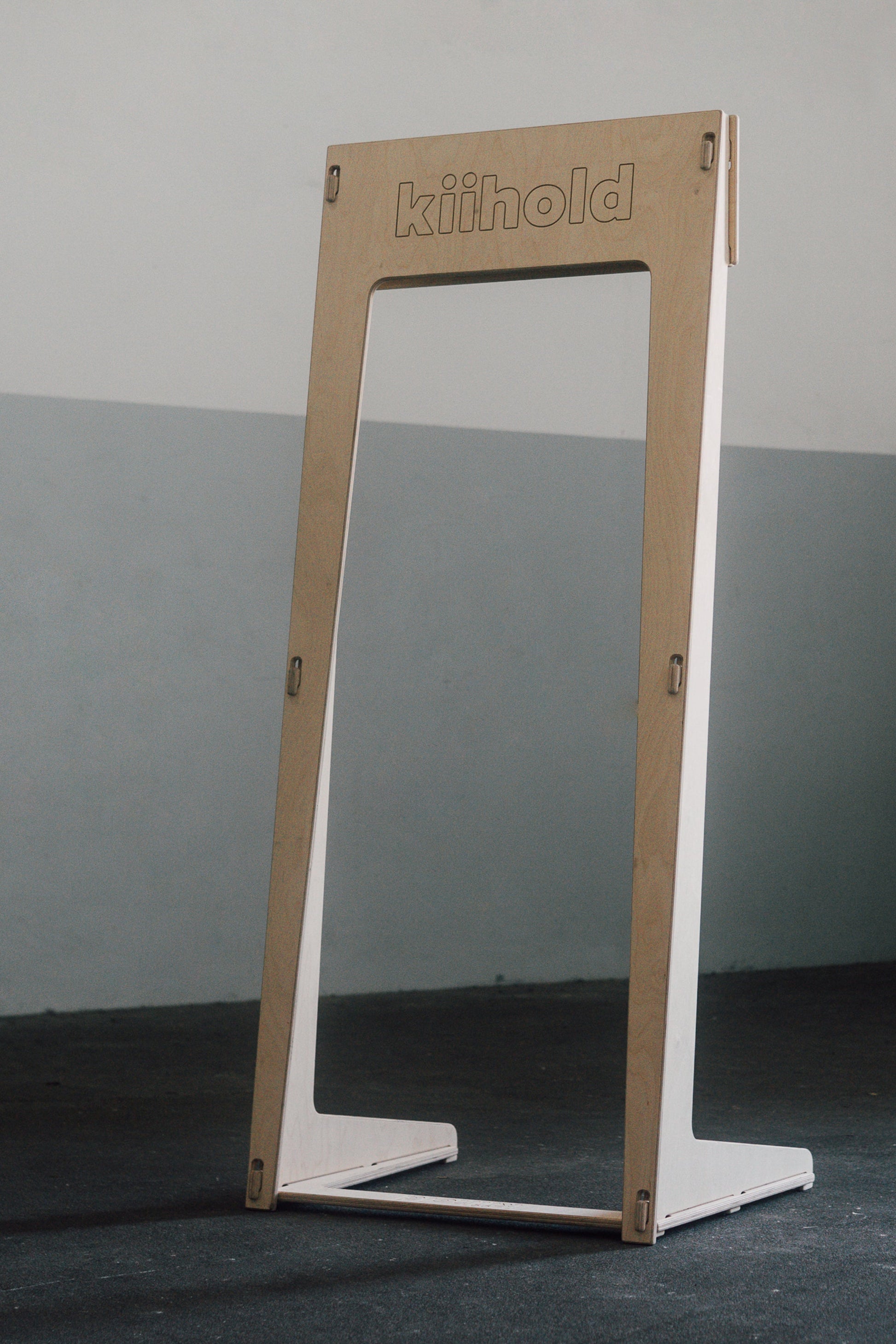 Freestanding wooden hanger with 'Kiihold' branding on a gray floor against a gray wall

